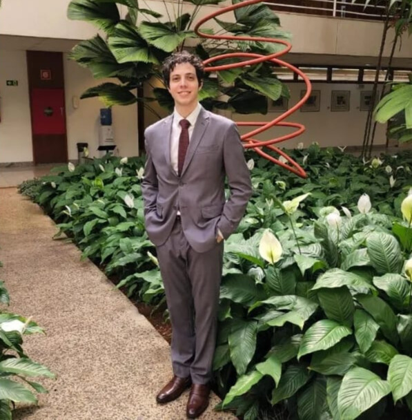 Homem branco com cabelos pretos, sorrindo, vestindo terno cinza, calça cinza, camisa branca e gravata vermelha. Ele está com as mãos nos bolsos da calça, parado em frente a um prédio com jardim e uma obra de arte vermelha.